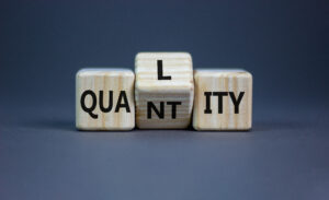 Wooden blocks spelling "QUALITY" with the middle block showing an "N," altering it to "QUANTITY.