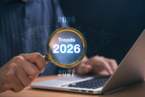 A person holds a magnifying glass showing "Trends 2026" in front of a laptop, with years 2025 and 2027 in the background over a world map graphic.