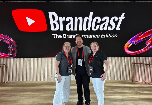 Three people stand and smile in front of a large Brandcast sign for "The Brand Performance Edition" event, with lanyards and badges visible.