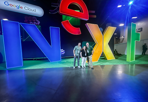 Three people stand in front of large, colorful letters spelling "Next" at a Google Cloud event indoors, with bright lights illuminating the scene.