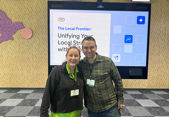 Two people stand and smile in front of a presentation screen displaying the title "Unifying Your Local Strategy with GPS.