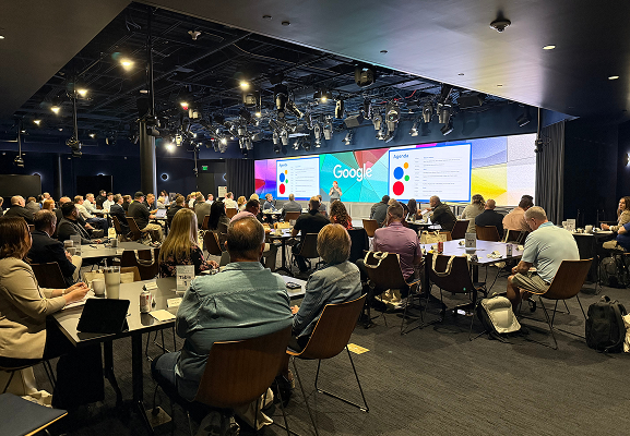 A large group of people sit at round tables in a conference room, facing a stage with Google branding and presentation slides displayed on large screens.